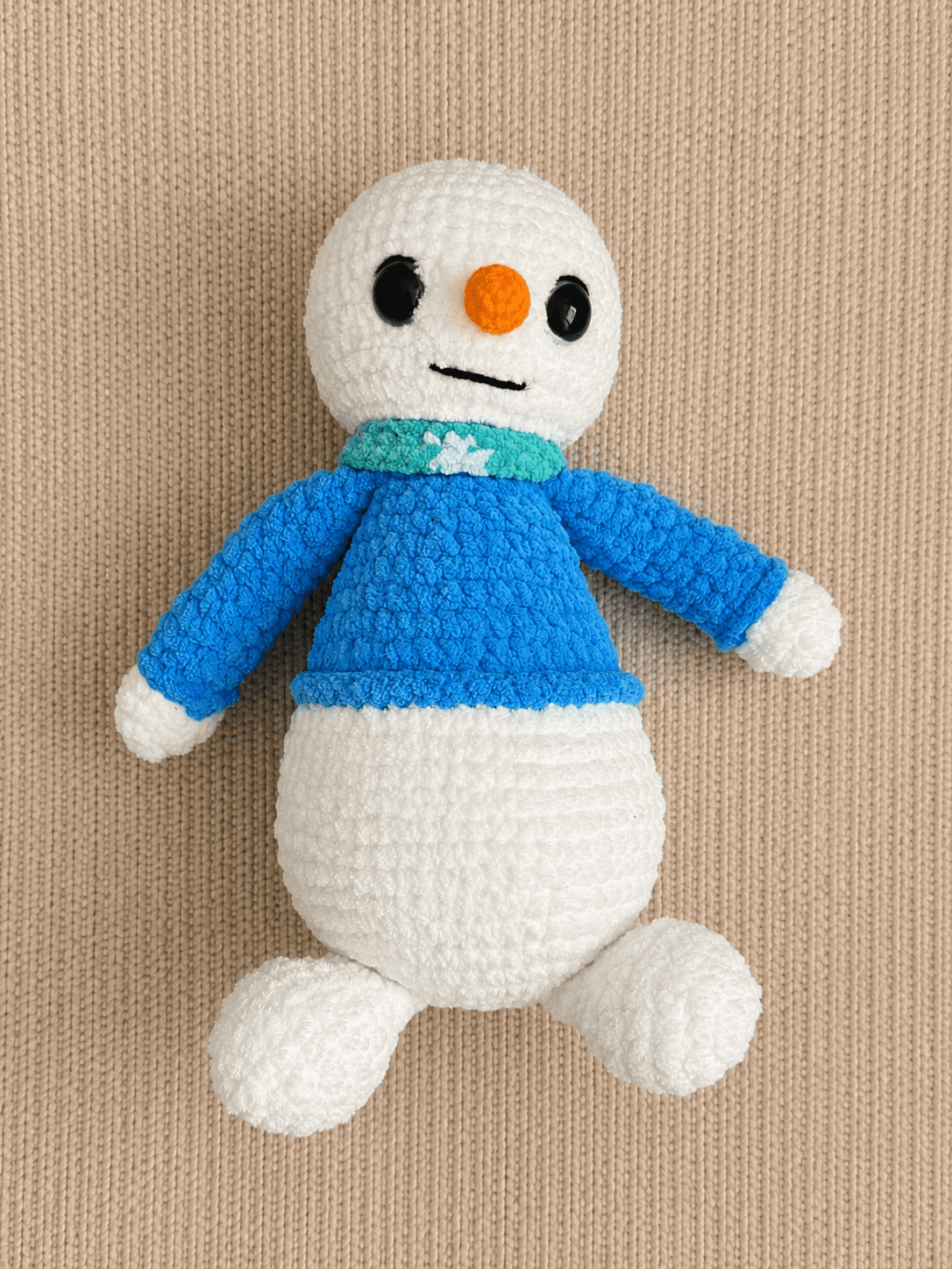 Top-down view of handmade crochet winter snowman plush laying on a textured cream background.