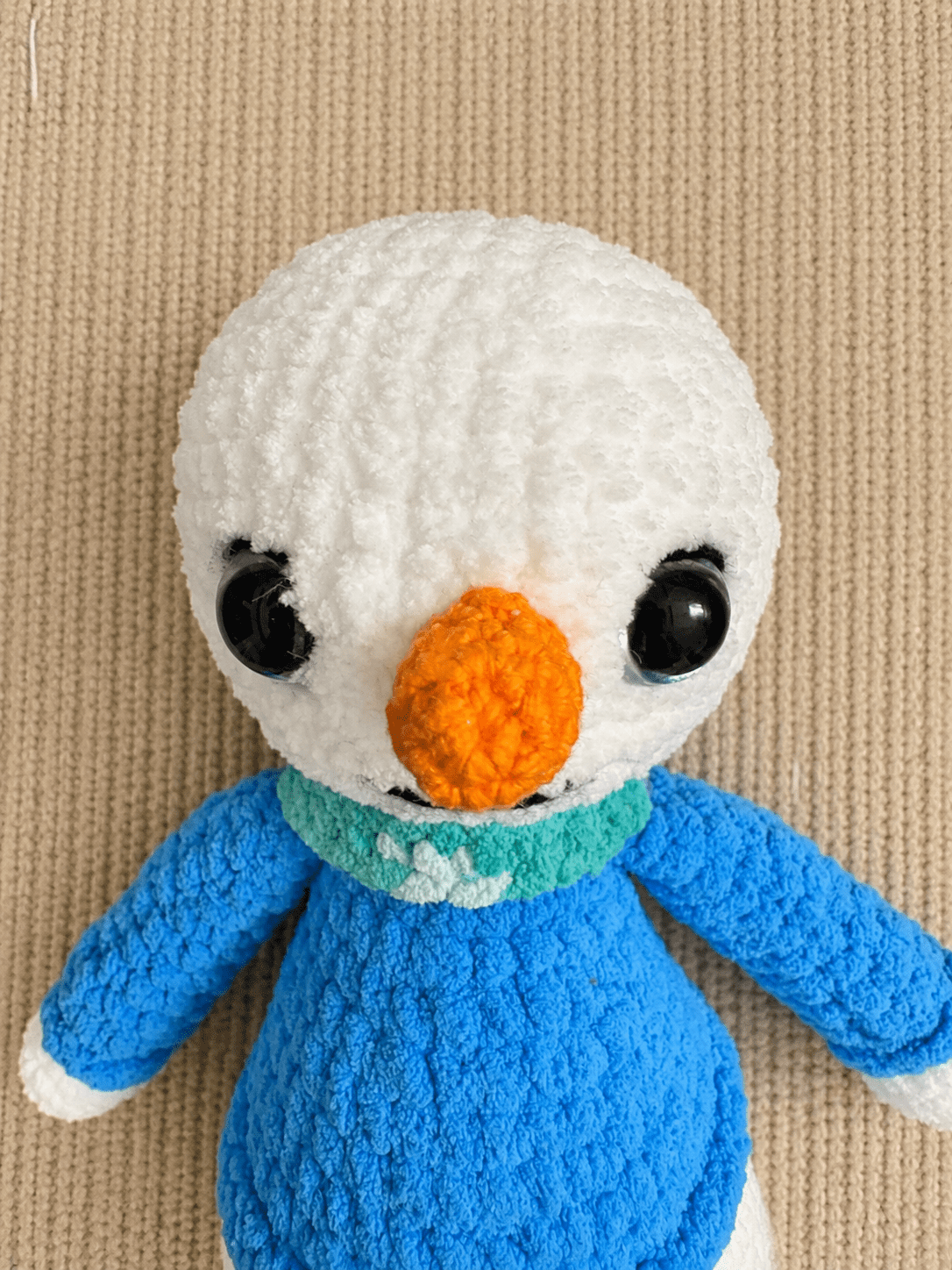 Close-up of handmade crochet snowman face with carrot nose and blue scarf.